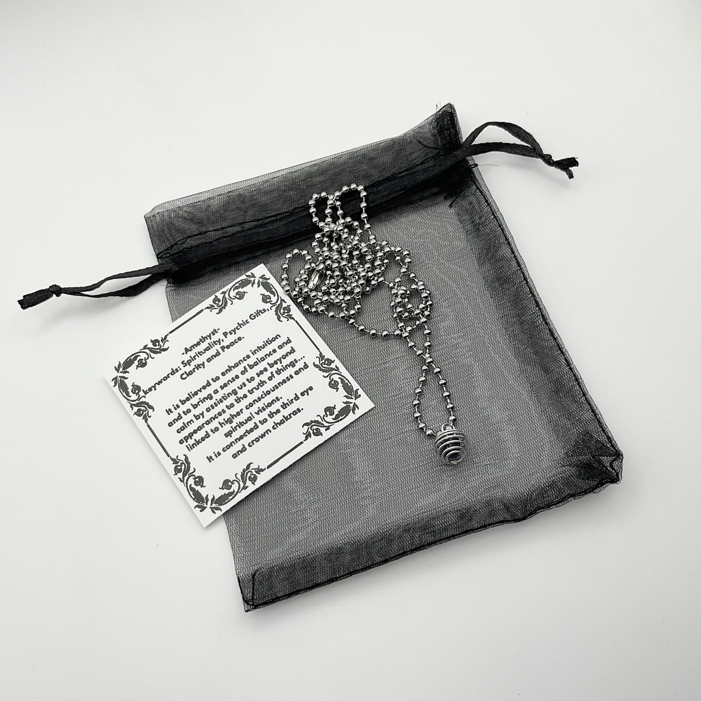 Ball Chain necklace with cage containing amethyst chip on a black drawstring organza bag on a light gray background