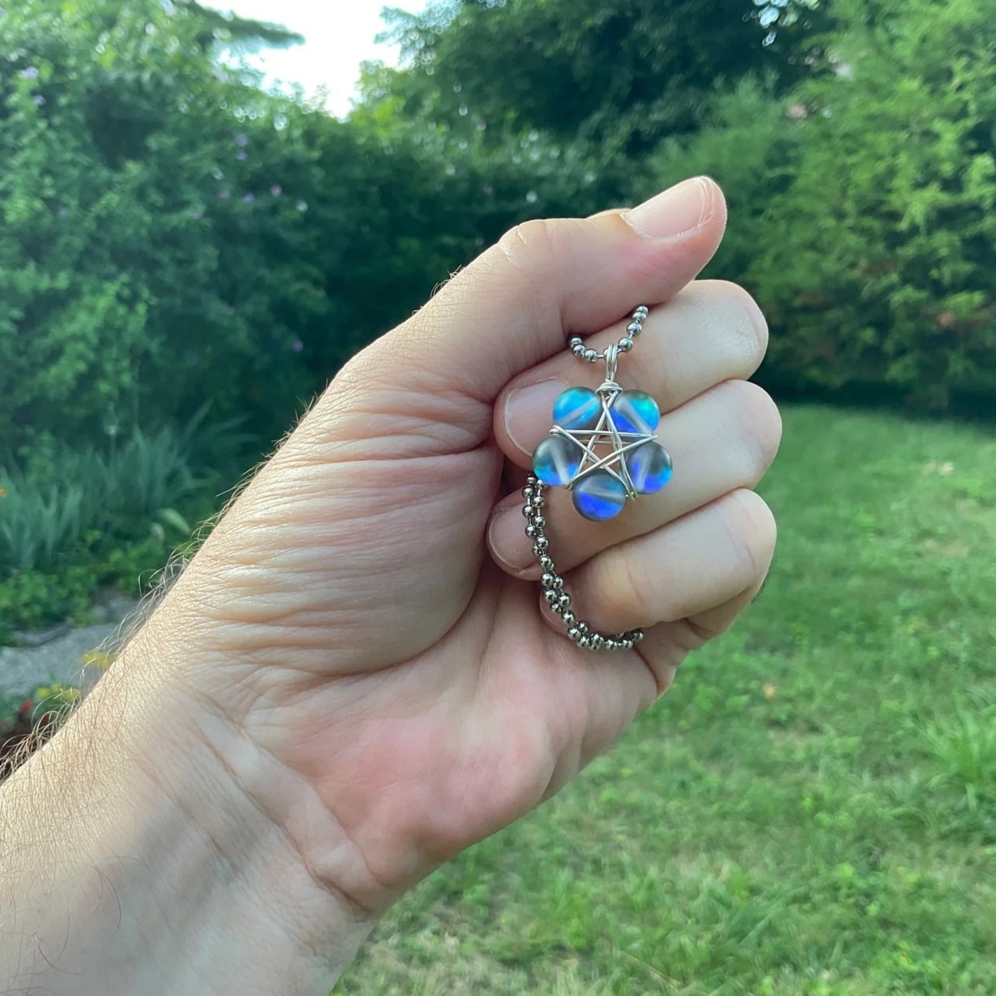 Hand holding a necklace with a blue tone pentagram wire wrap bead pendant against a green garden outdoor background