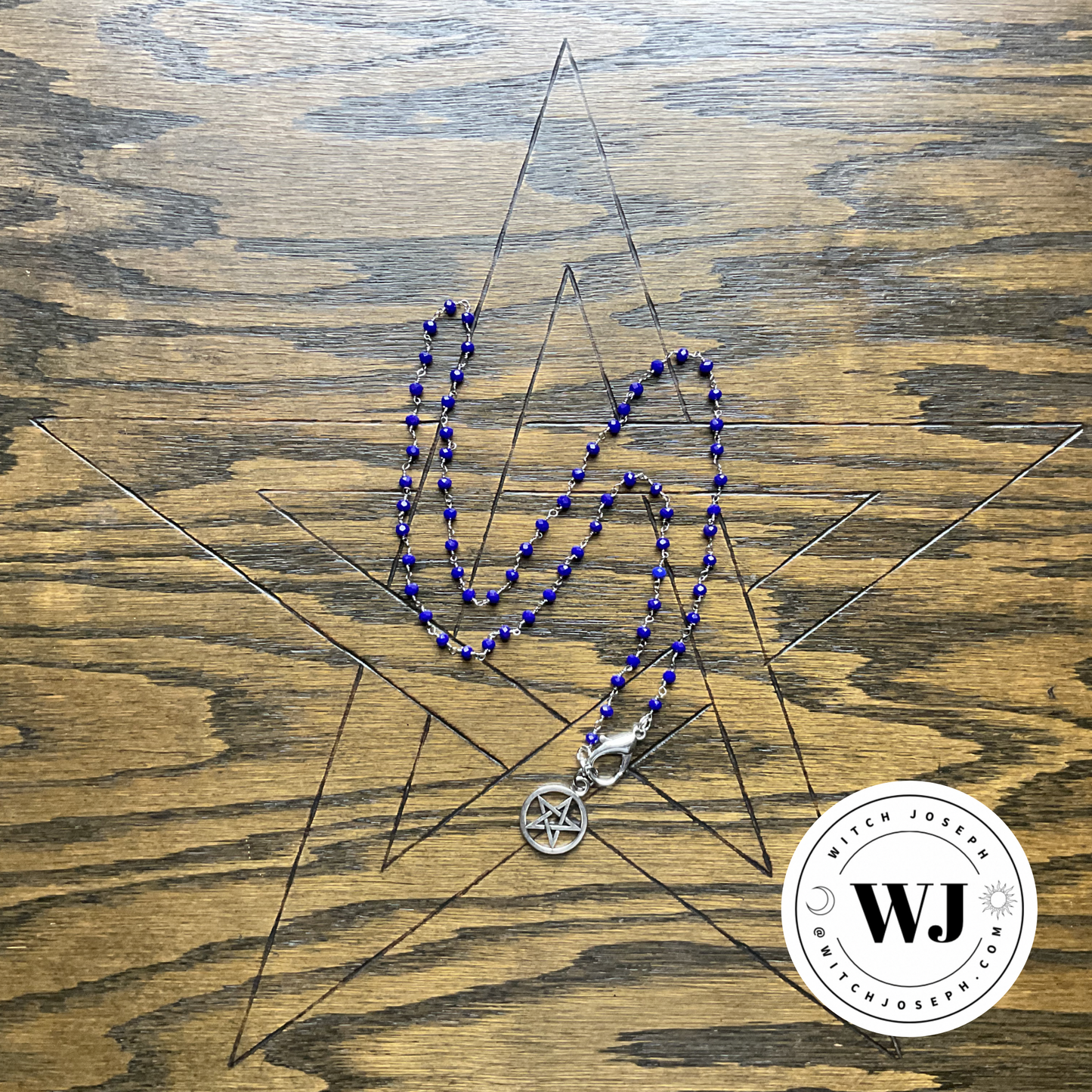Blue beads on silver tone necklace with a silver tone pentacle pendant displayed on a wooden surface, featuring the Witch Joseph logo.