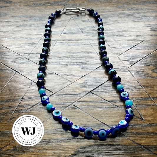 A beaded necklace with evil eye charms in shades of blue, black, and turquoise, displayed on a dark wood background. A circular 'Witch Joseph' watermark is visible in the bottom-left corner.