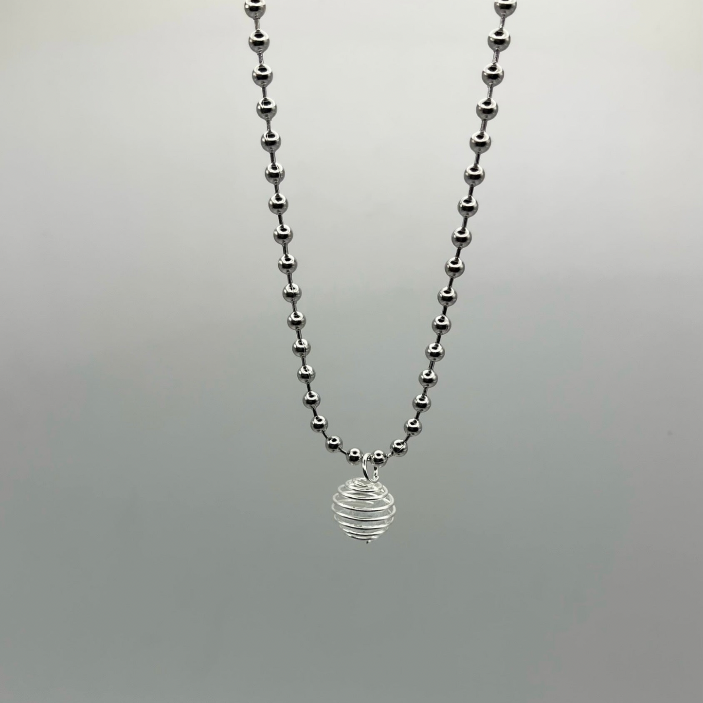 Silver tone ball chain necklace with a small round silver tone cage pendant containing a celestite crystal chip laying on a light gray background