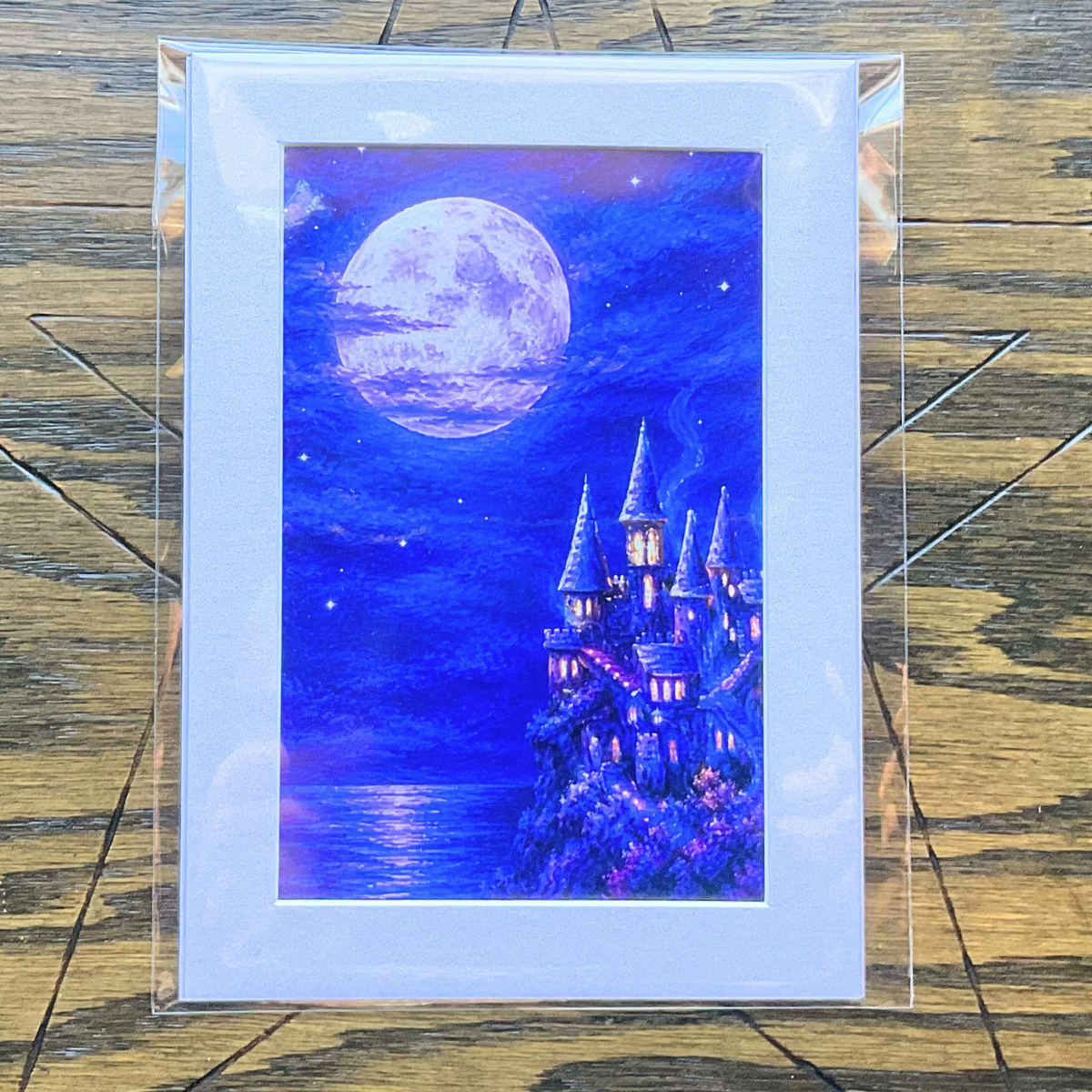 A matted art print depicting a whimsical purple and blue castle under a large glowing moon, mounted in a clear protective sleeve and displayed on a rustic wooden background.