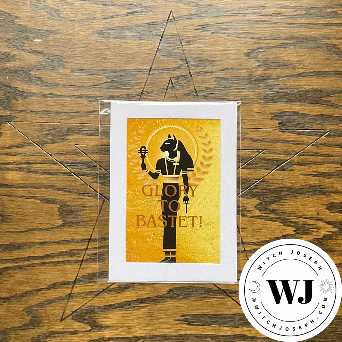 An art print featuring a black silhouette of Bastet on a textured gold background with the text 'GLORY TO BASTET!' in gold, displayed on a wooden surface inside a clear acrylic package. A circular 'Witch Joseph' brand logo is in the bottom right corner.