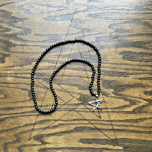 Black beaded necklace with a silver tone inverted pentagram pendant on a wooden surface with the pentagram etched into it