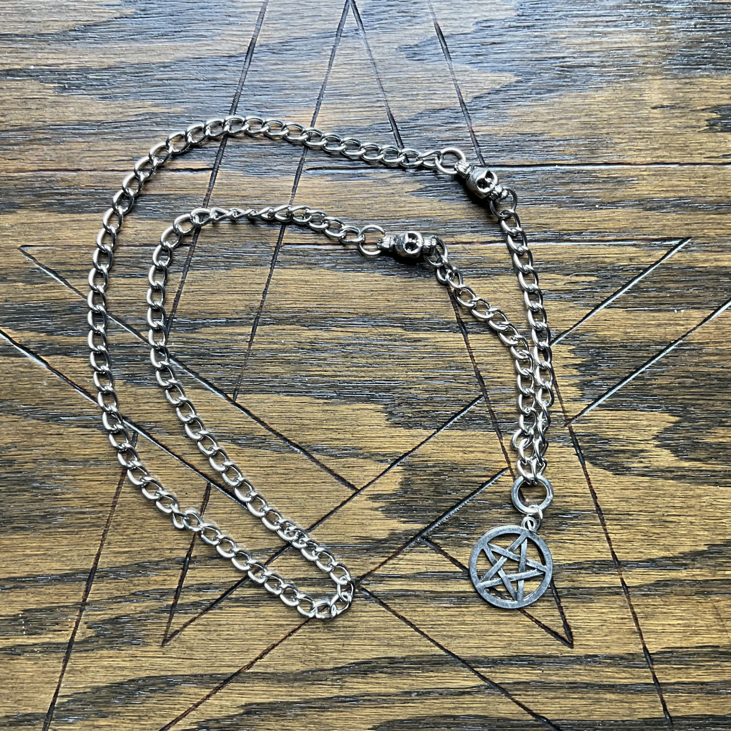 Silver tone chain necklace with a pentacle pendant with skull accent beads displayed on a wooden surface with an etched pentagram in it