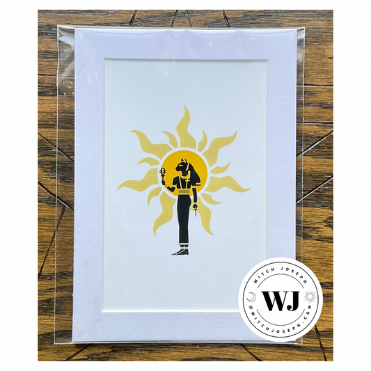 an art print of the Goddess Bastet carrying sistrum and ankh against a solar image with white mat displayed against a wooden background with the Witch Joseph Logo shown in the lower right corner