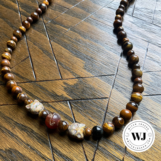 A beaded necklace made of polished brown tiger's eye beads, featuring three unique carved accent beads, displayed on a dark wood parquet floor. A circular 'Witch Joseph' logo is visible in the bottom right corner.