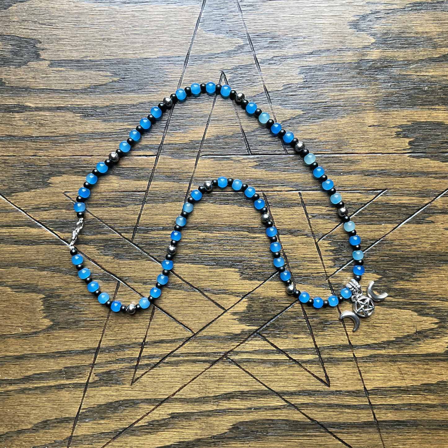 Necklace with blue and black beads with a silver tone triple goddess charm displayed on a wooden surface with a pentagram etched into it