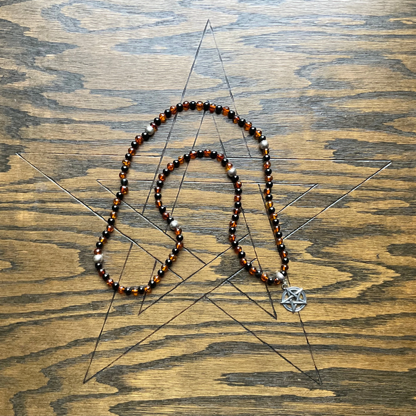 Amber and Jet Beaded necklace with a Sterling Silver pentagram pendant and sterling silver accent beads displayed on a wooden surface with an etched Pentagram.