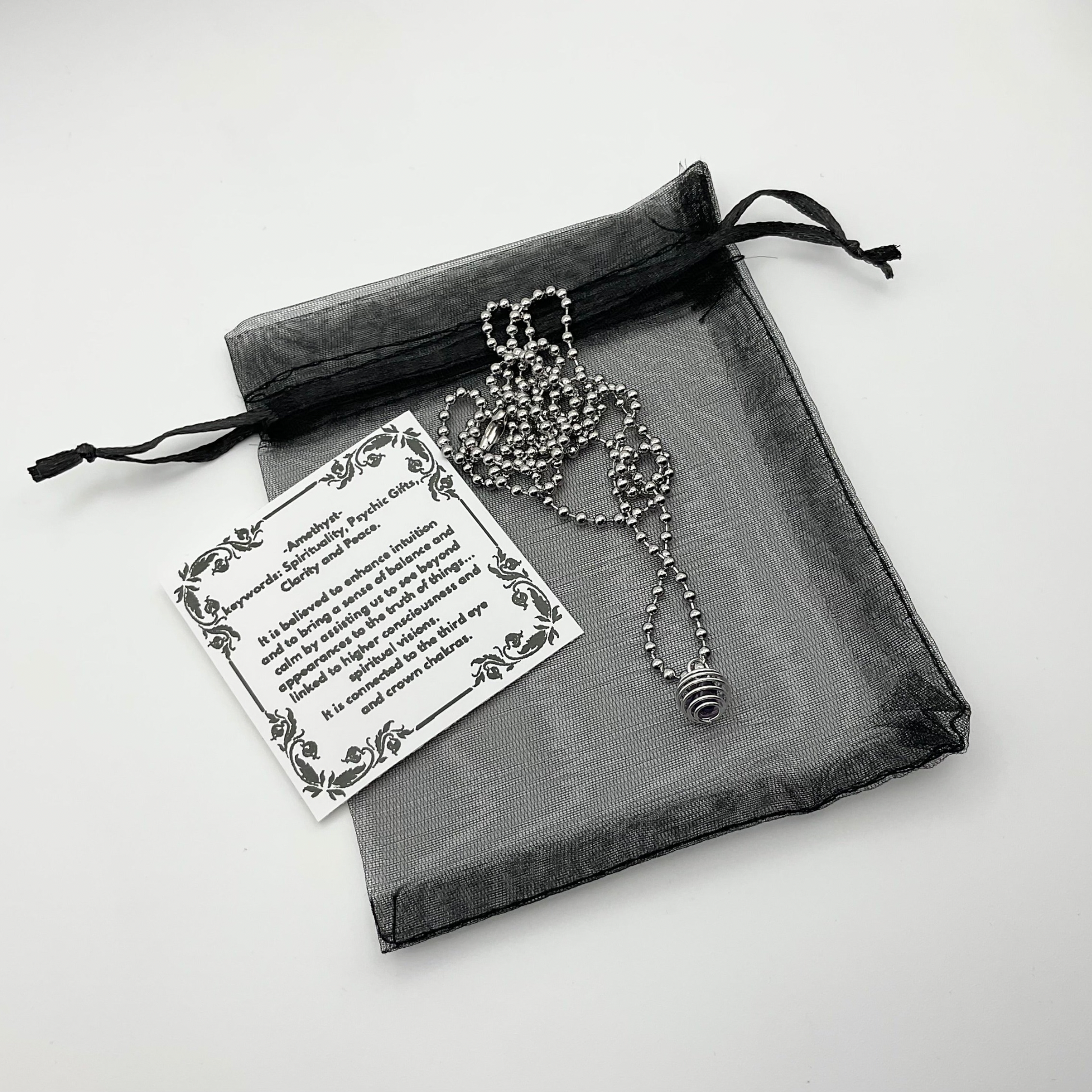 Ball Chain necklace with cage containing amethyst chip on a black drawstring organza bag on a light gray background
