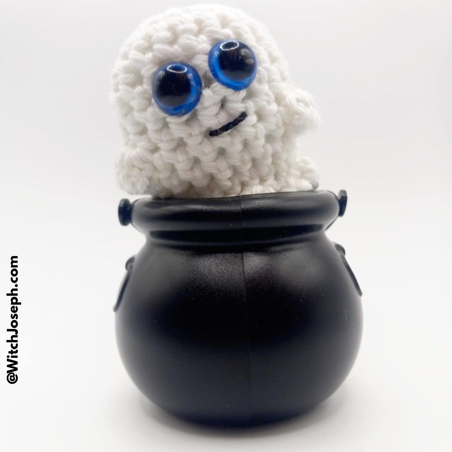 White Amigurumi Ghost glows in dark with blue eyes inside a black cauldron on a white background with the Witch Joseph URL