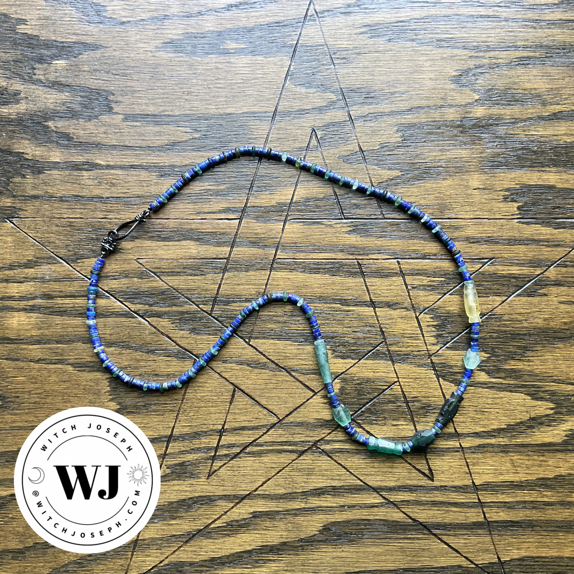 Necklace with Roman glass and lapis lazuli beads in blue and green beads displayed on a wooden surface with a Pentagram design, featuring the Witch Joseph logo