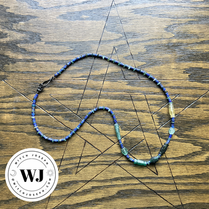 Necklace with Roman glass and lapis lazuli beads in blue and green beads displayed on a wooden surface with a Pentagram design, featuring the Witch Joseph logo