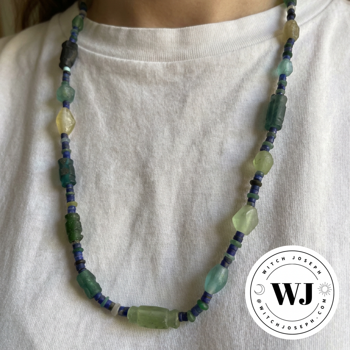 Necklace with ancient Roman glass green and blue beads with lapis beads displayed on a model in a white shirt, featuring the Witch Joseph logo