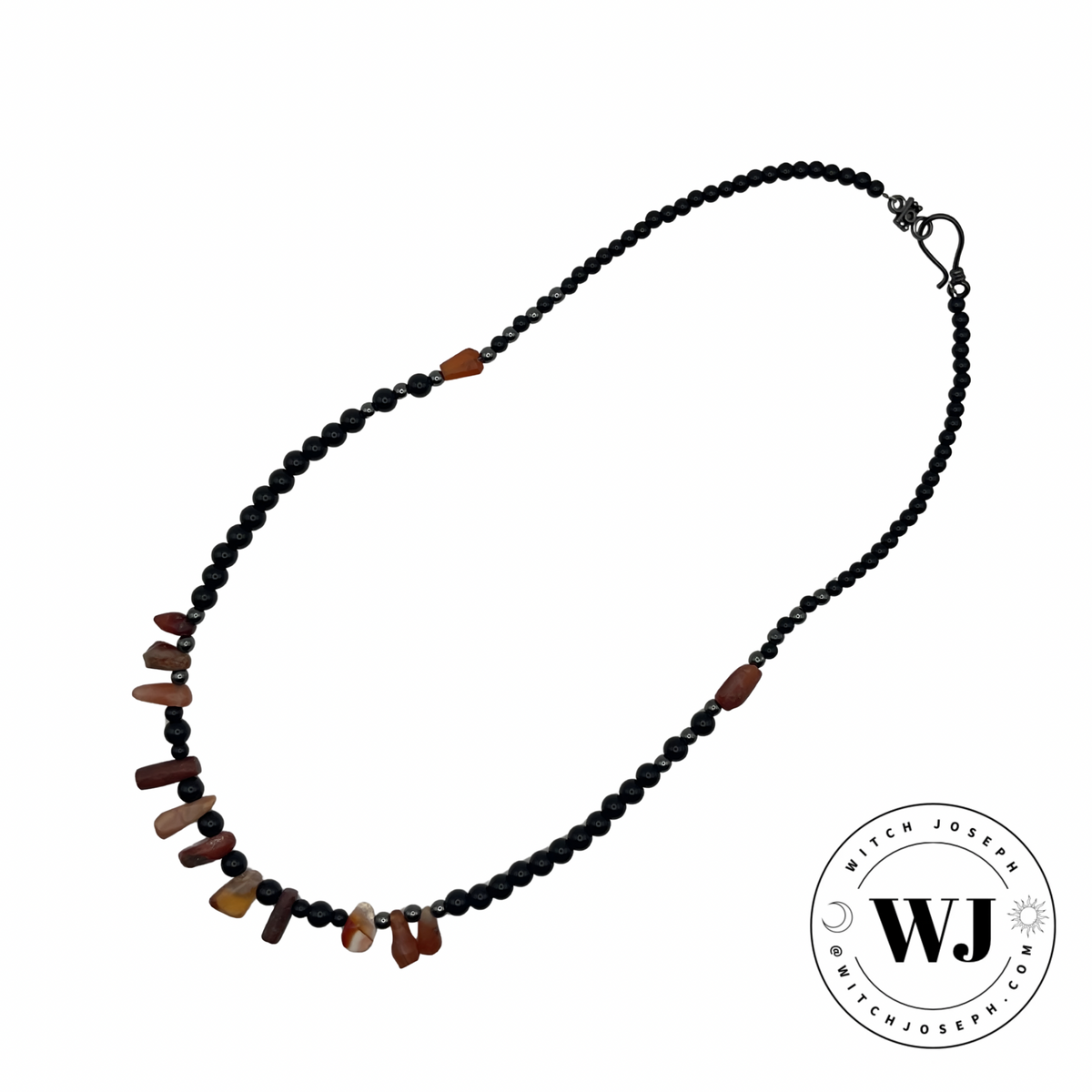A beaded necklace featuring a strand of small black beads and small hematite accent beads with an antique carnelian beads arranged periodically through the necklace, shown on a plain white background with a circular witch joseph logo in the bottom right corner.