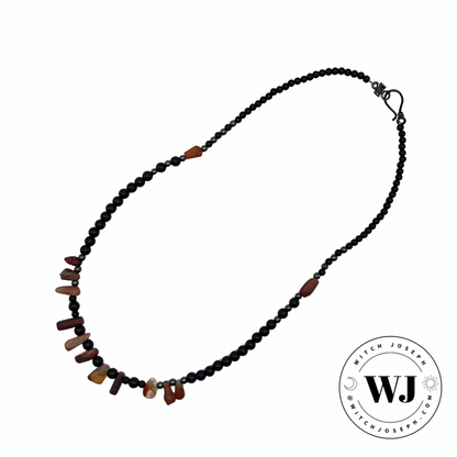 A beaded necklace featuring a strand of small black beads and small hematite accent beads with an antique carnelian beads arranged periodically through the necklace, shown on a plain white background with a circular witch joseph logo in the bottom right corner.