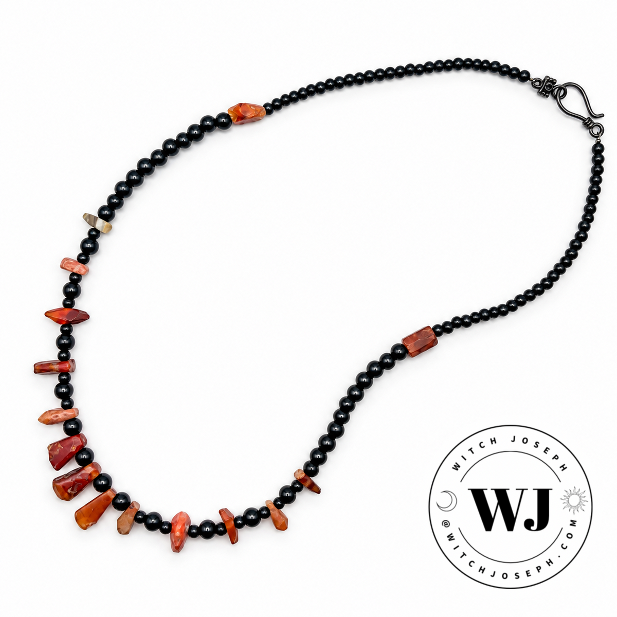 A beaded necklace featuring black round beads with irregular, polished red and orange antique carnelian beads spaced along the front, displayed on a clean white background with a circular 'Witch Joseph' logo in the lower right corner.
