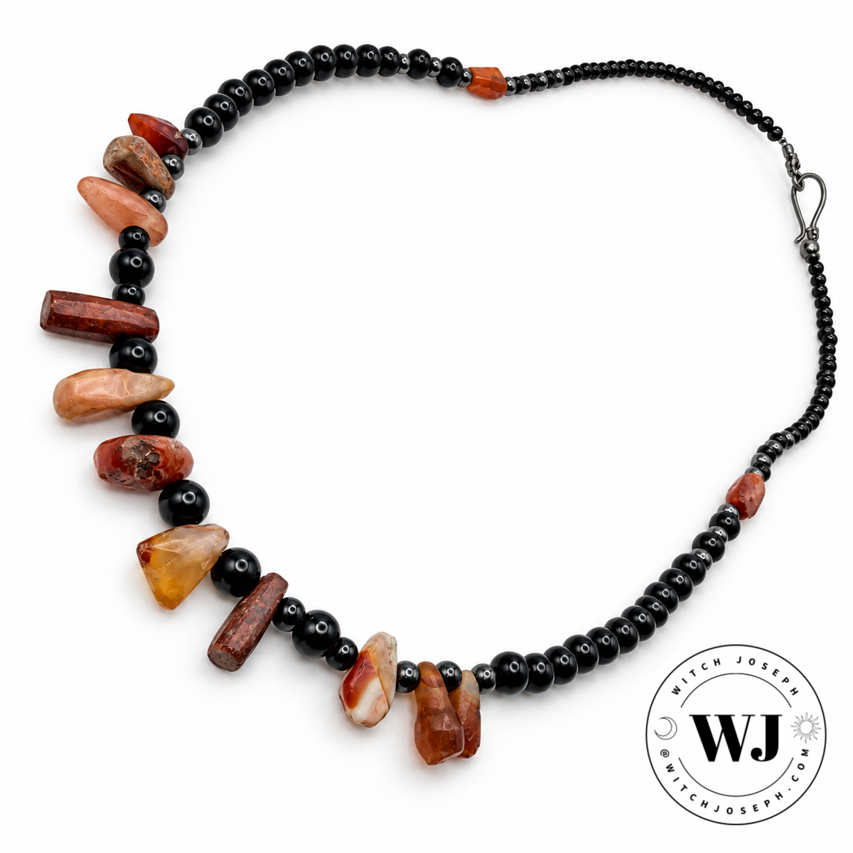 A beaded necklace featuring black beads and hematite beads and irregular polished antique carnelian chalcedony beads in earthy terracotta, reddish-brown, and amber tones, displayed on a clean white background with a circular 'Witch Joseph' Logo in the lower right corner.