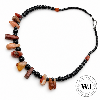 A beaded necklace featuring black beads and hematite beads and irregular polished antique carnelian chalcedony beads in earthy terracotta, reddish-brown, and amber tones, displayed on a clean white background with a circular 'Witch Joseph' Logo in the lower right corner.