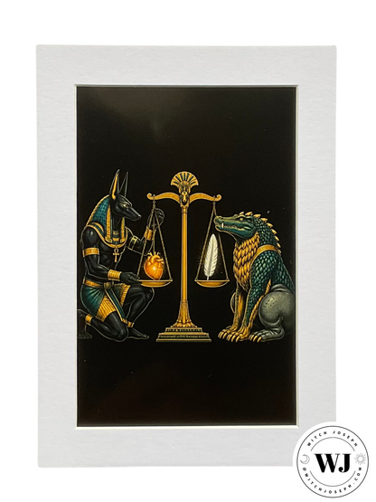 A matted art print featuring an Egyptian-themed illustration of Anubis and Ammit with golden scales on a black background, set within a wide white Mat.  They are shown weighing the heart against the feather of Maat.  The witch joseph logo is shown in the lower right hand corner.