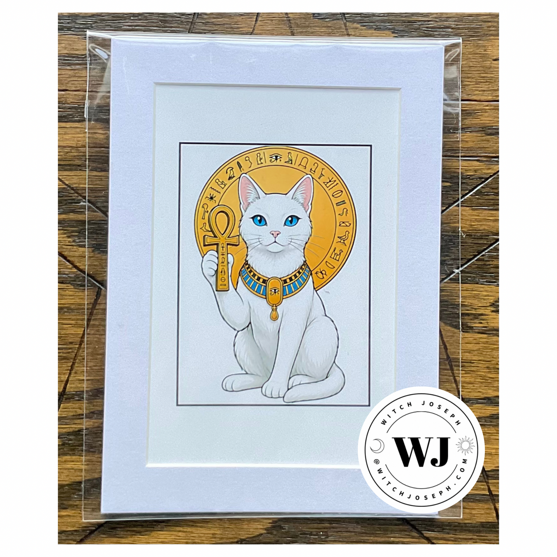 A framed art print featuring a white cat with blue eyes holding an ankh inside a gold circular emblem, presented in a white mat and clear plastic sleeve on a wooden surface. A circular 'Witch Joseph' logo brand watermark is visible in the bottom right corner.