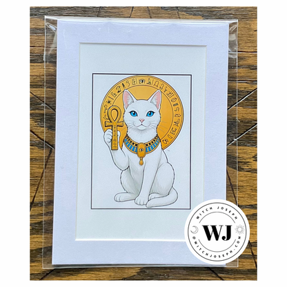 A framed art print featuring a white cat with blue eyes holding an ankh inside a gold circular emblem, presented in a white mat and clear plastic sleeve on a wooden surface. A circular 'Witch Joseph' logo brand watermark is visible in the bottom right corner.