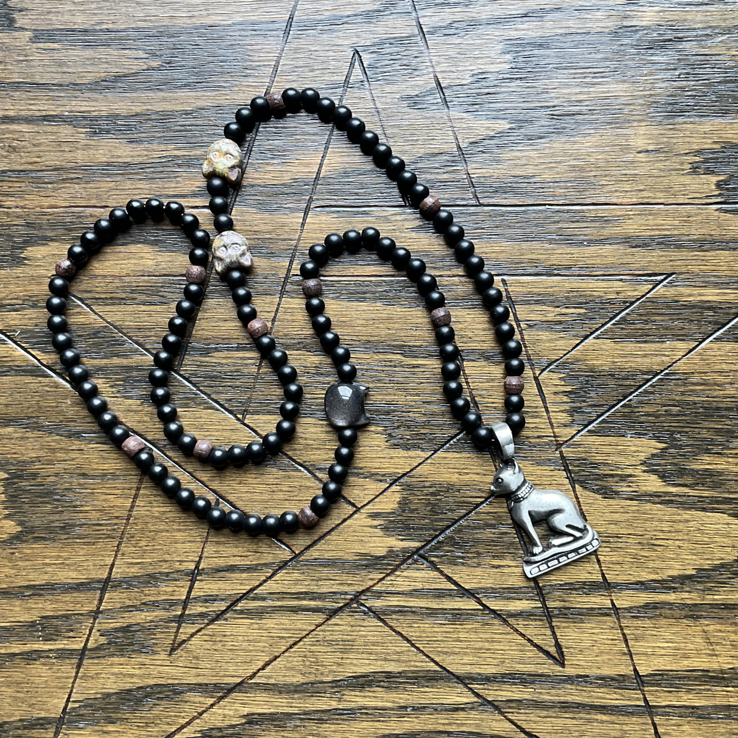 Black beaded Bastet necklace with a silver tone Bastet  pendant displayed on a wooden surface with a pentagram etched in to it
