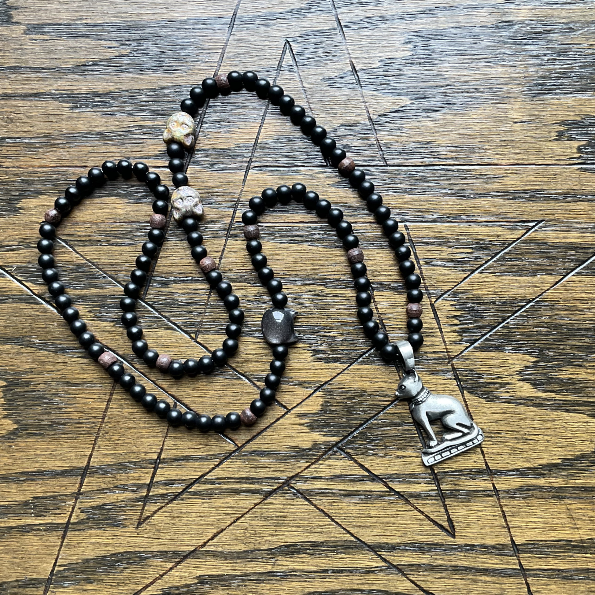 Black beaded Bastet necklace with a silver tone Bastet  pendant displayed on a wooden surface with a pentagram etched in to it