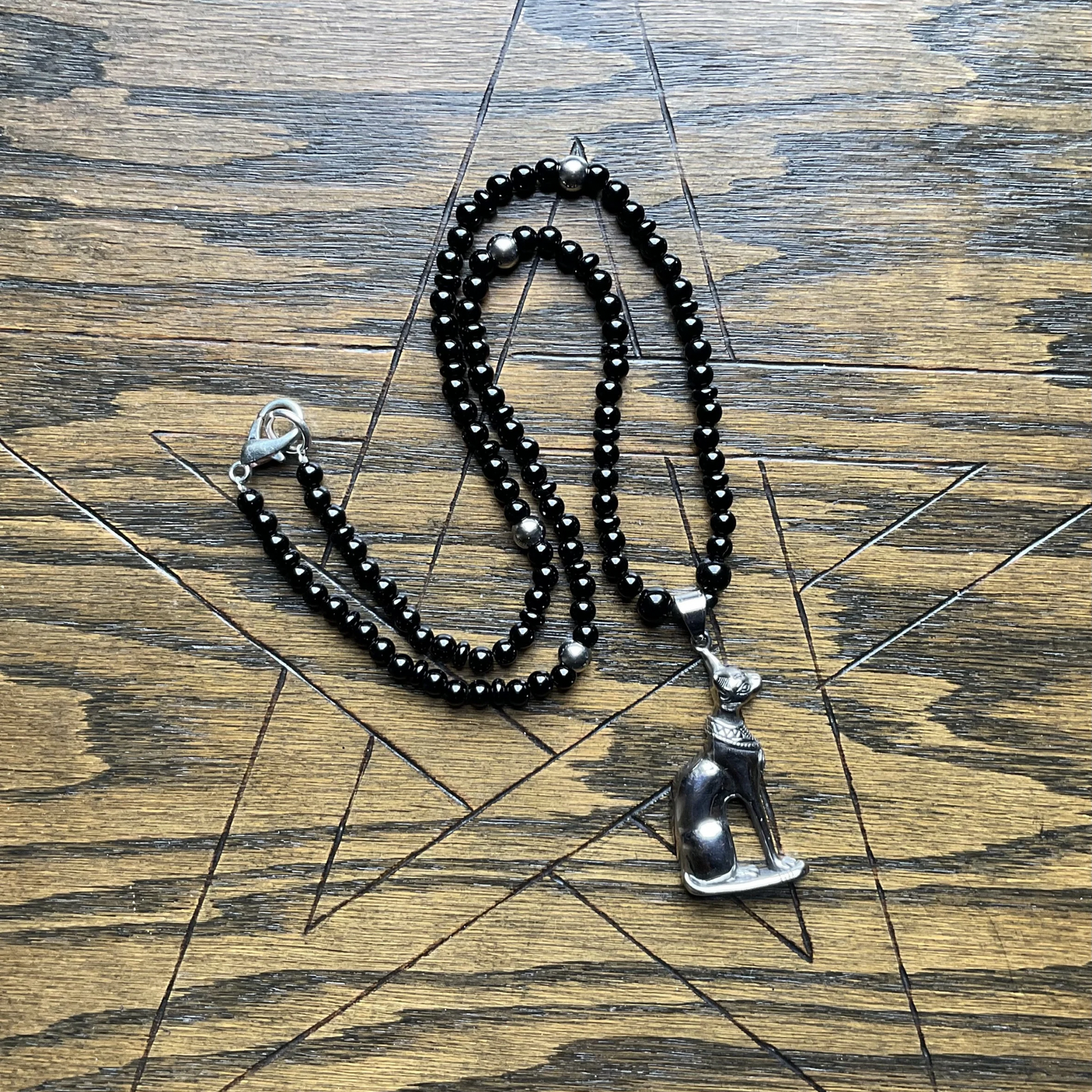 Black beaded necklace with silver tone accents with a silver tone Cat Goddess pendant on a wooden surface with a pentagram etched into it