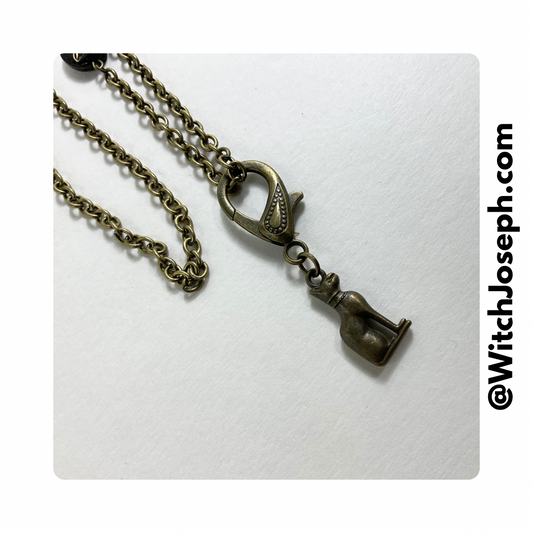 A bronze-toned necklace with a cat-shaped pendant lobster claw and a detachable chain. The brand's website is visible in the corner.