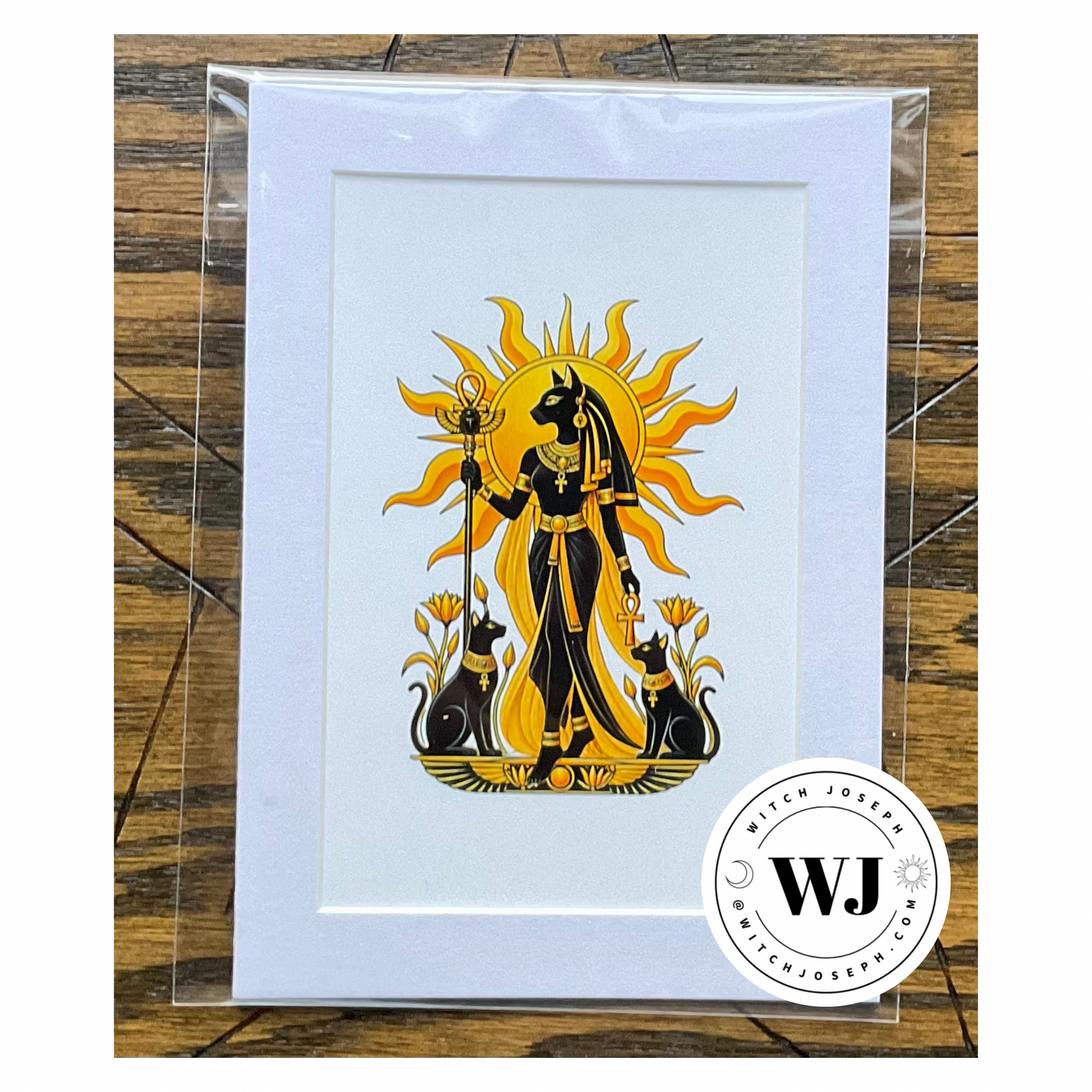 A matted art print featuring a stylized black and gold illustration of Bastet with Her sacred cats, sealed in a clear plastic sleeve and photographed on a wooden surface. A circular 'Witch Joseph' logo brand is visible in the bottom right corner.