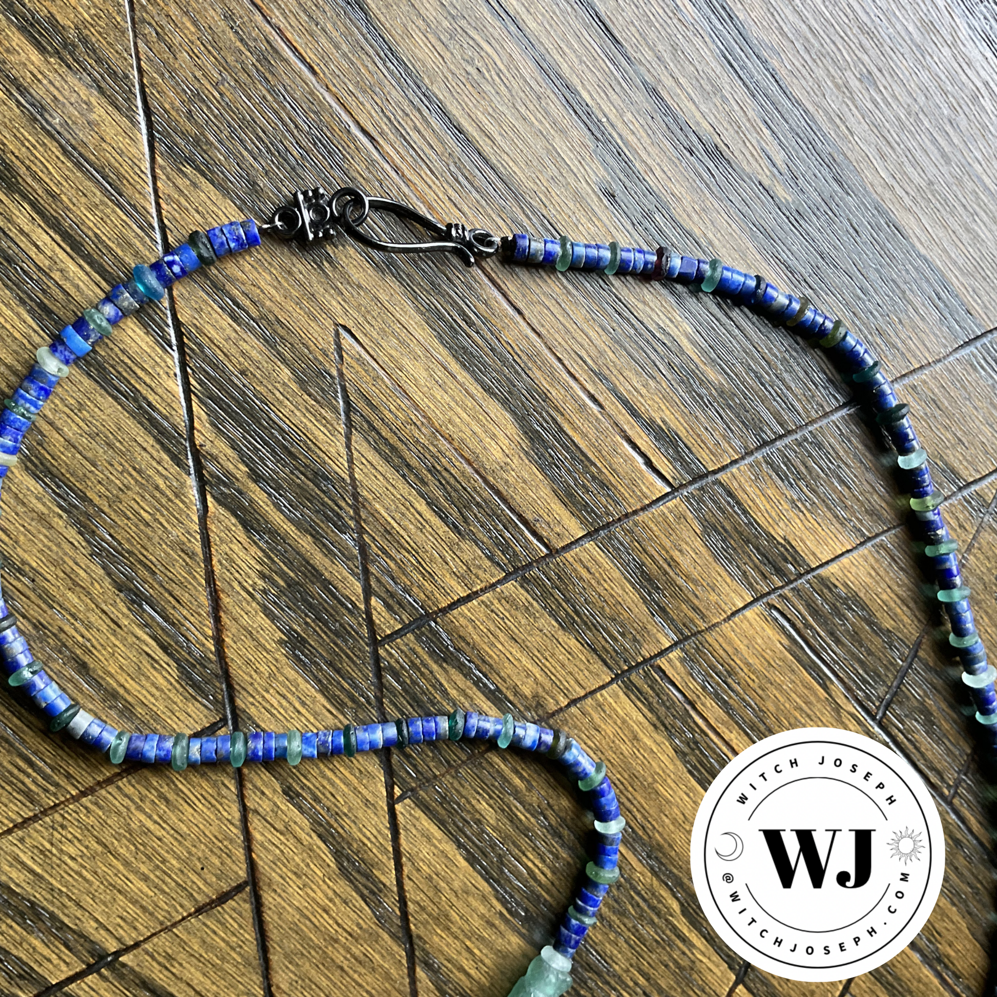Roman Glass and Lapis Blue beaded necklace with gun metal tone metal clasp displayed on a wooden surface with a pentagram etched into it featuring the Witch Joseph logo.