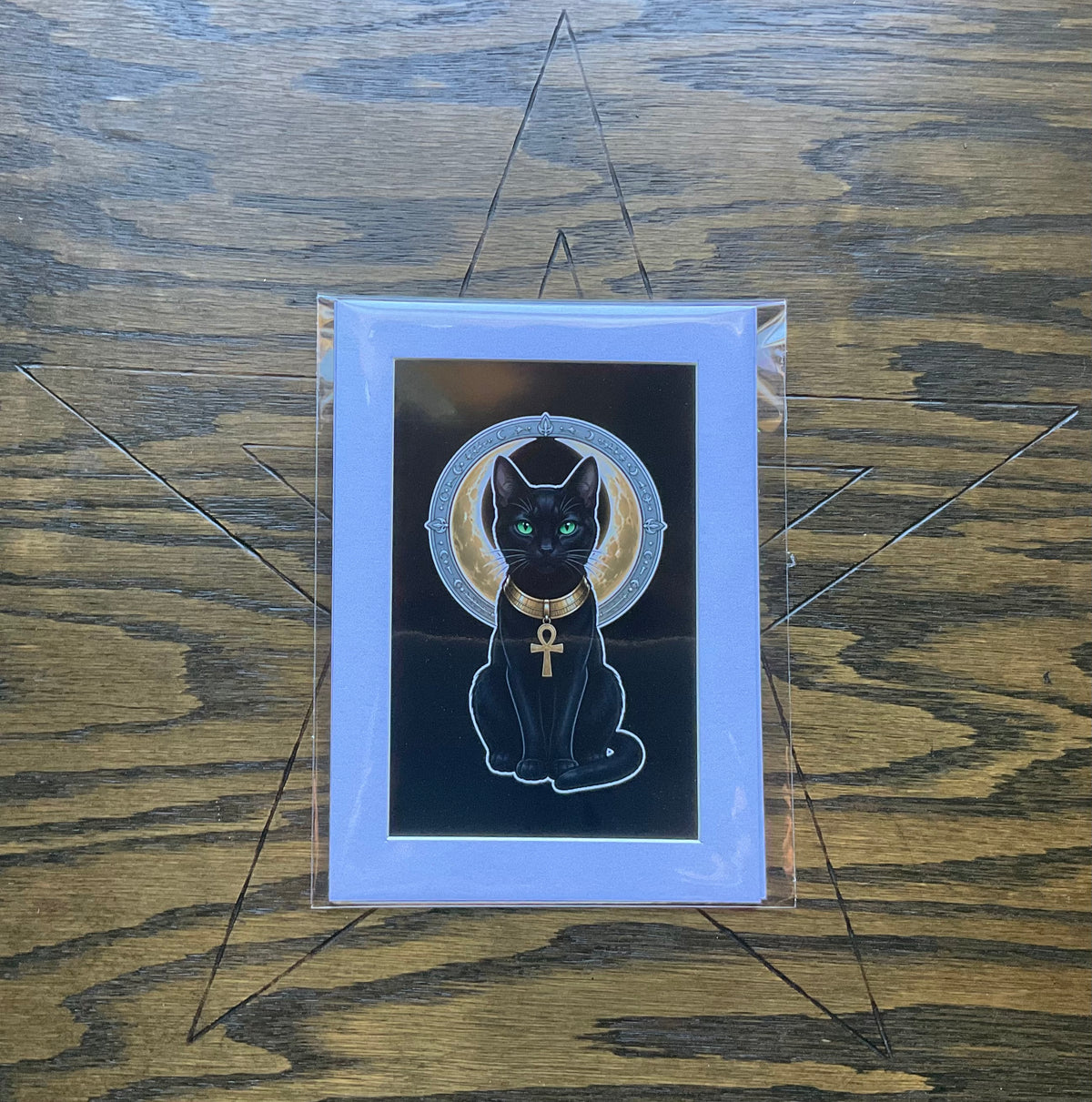A art print featuring a black cat Bast with green eyes, wearing a gold collar with an ankh symbol and set against a gold halo on a black background, mounted on a white mat and enclosed in a protective sleeve. The print is displayed on a dark wood background.