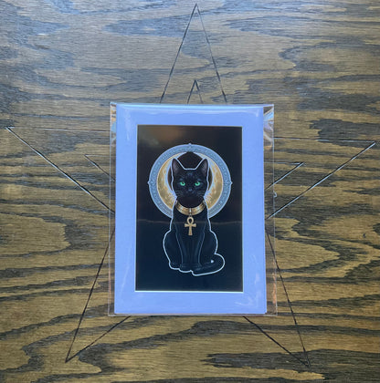 A art print featuring a black cat Bast with green eyes, wearing a gold collar with an ankh symbol and set against a gold halo on a black background, mounted on a white mat and enclosed in a protective sleeve. The print is displayed on a dark wood background.