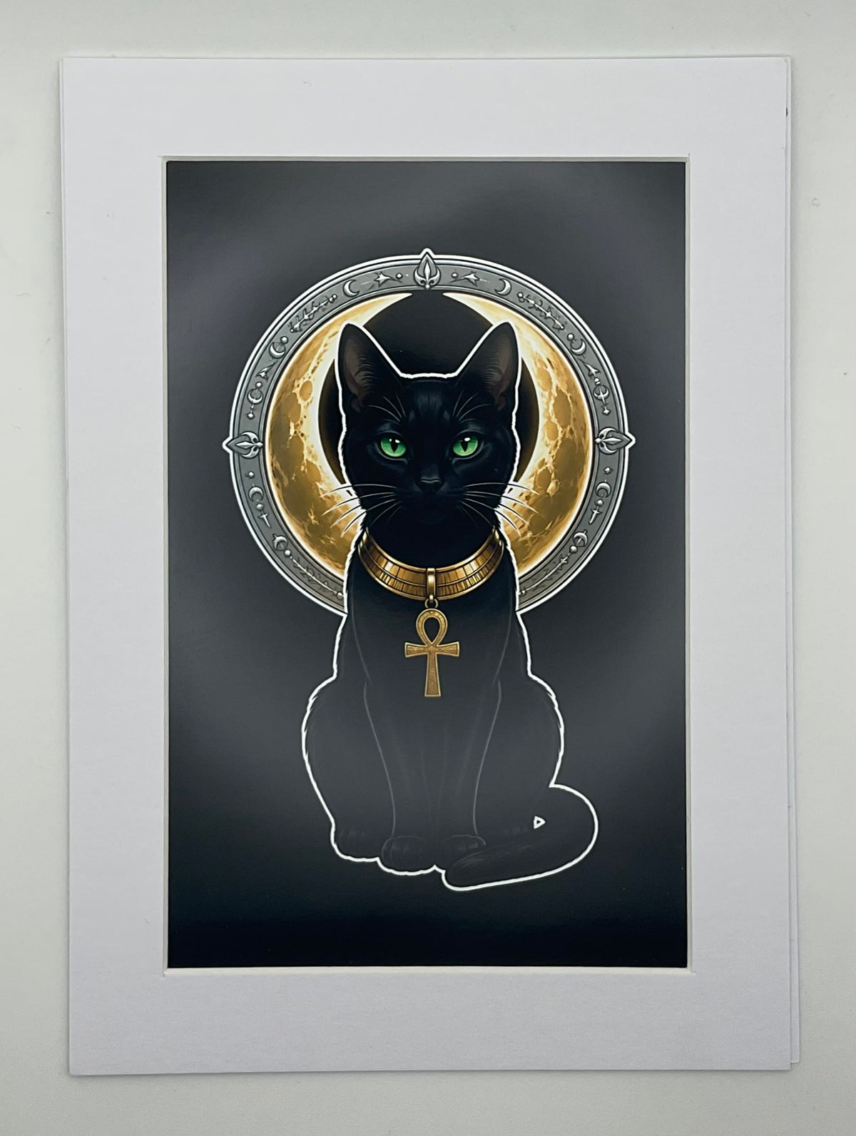 A matted art print featuring a black cat Bastet with glowing green eyes, wearing a gold collar with an ankh pendant, set against a dark background with a circular gold and silver halo motif. The print is mounted in a white Mat.