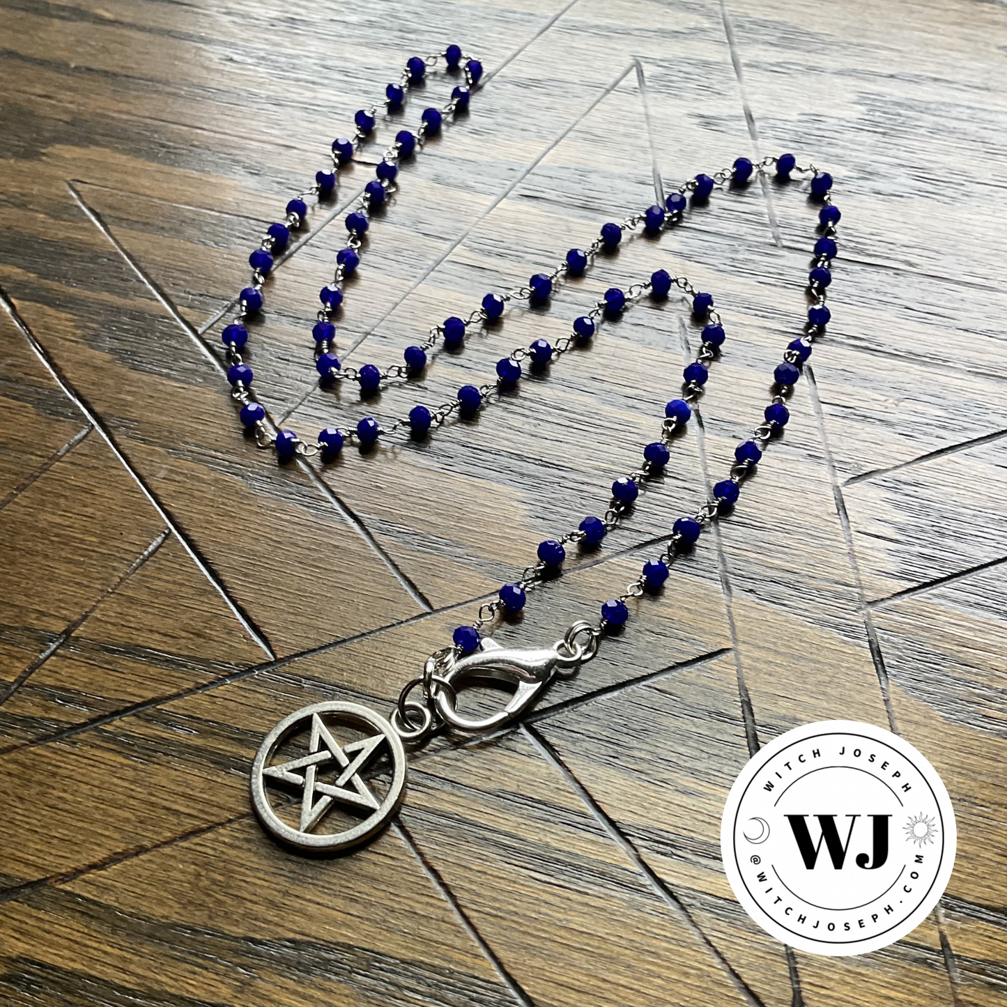 Necklace with blue beads and silver tone pentacle pendant displayed on a wooden surface with an etched pentagram, featuring the Witch Joseph Logo.