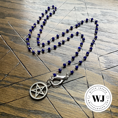Necklace with blue beads and silver tone pentacle pendant displayed on a wooden surface with an etched pentagram, featuring the Witch Joseph Logo.