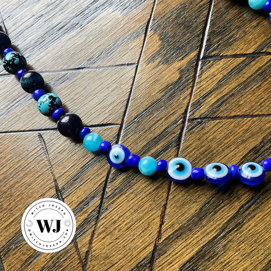 A beaded necklace featuring evil eye beads and blue tigers eye with blue art and seed  bead accents, displayed on a dark wood background. A circular 'Witch Joseph' logo watermark is visible in the bottom-left corner.
