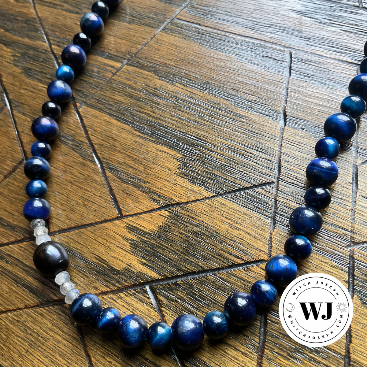 A beaded necklace made of polished dark blue tigers eye beads with a few small labradorite and black accent beads, displayed on a dark wood surface. A circular Witch Joseph logo is visible in the bottom right corner.