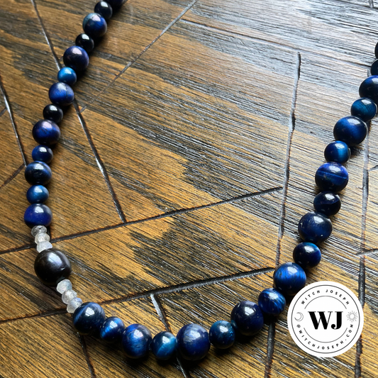 A beaded necklace made of polished dark blue tigers eye beads with a few small labradorite and black accent beads, displayed on a dark wood surface. A circular Witch Joseph logo is visible in the bottom right corner.