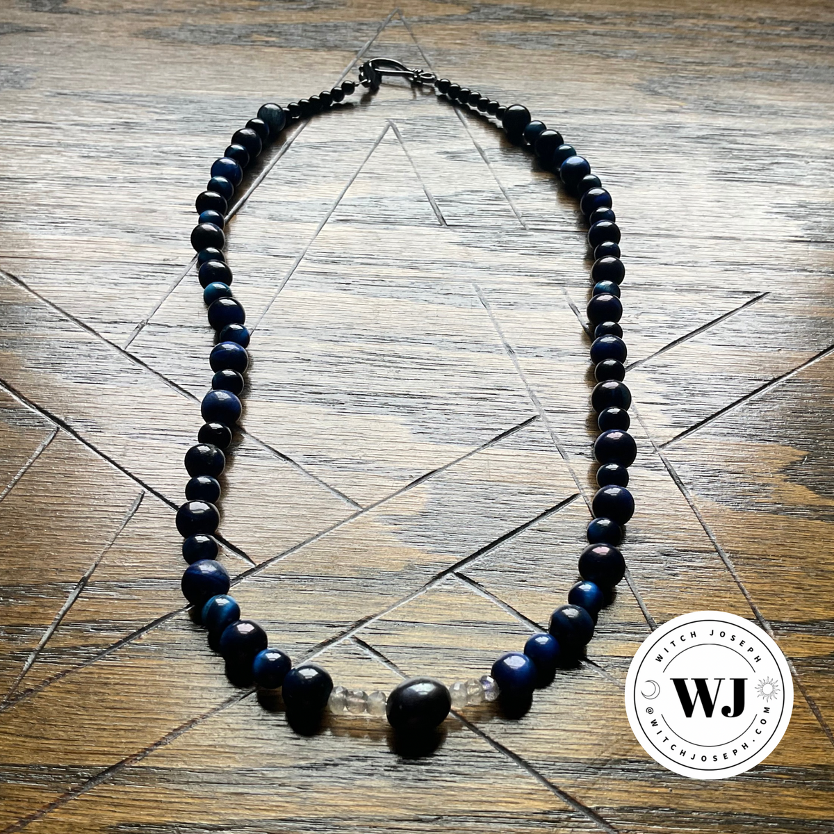 A beaded necklace with dark blue tigers eye and black round beads, featuring a central black pearl bead with labradorite chips…displayed on a wooden surface with a pentagram geometric pattern overlay. A circular 'Witch Joseph' watermark is in the bottom right corner.