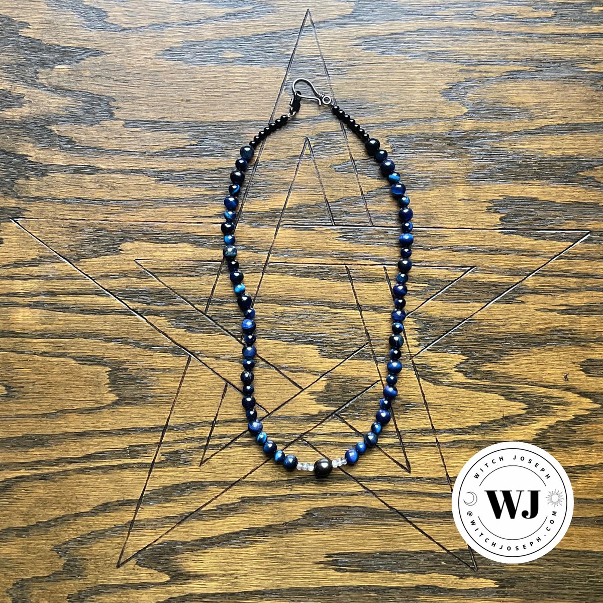 A beaded necklace with dark blue tiger eye and black beads, displayed on a wooden surface etched with a pentagram. A circular logo for 'Witch Joseph' logo is visible in the bottom right corner.