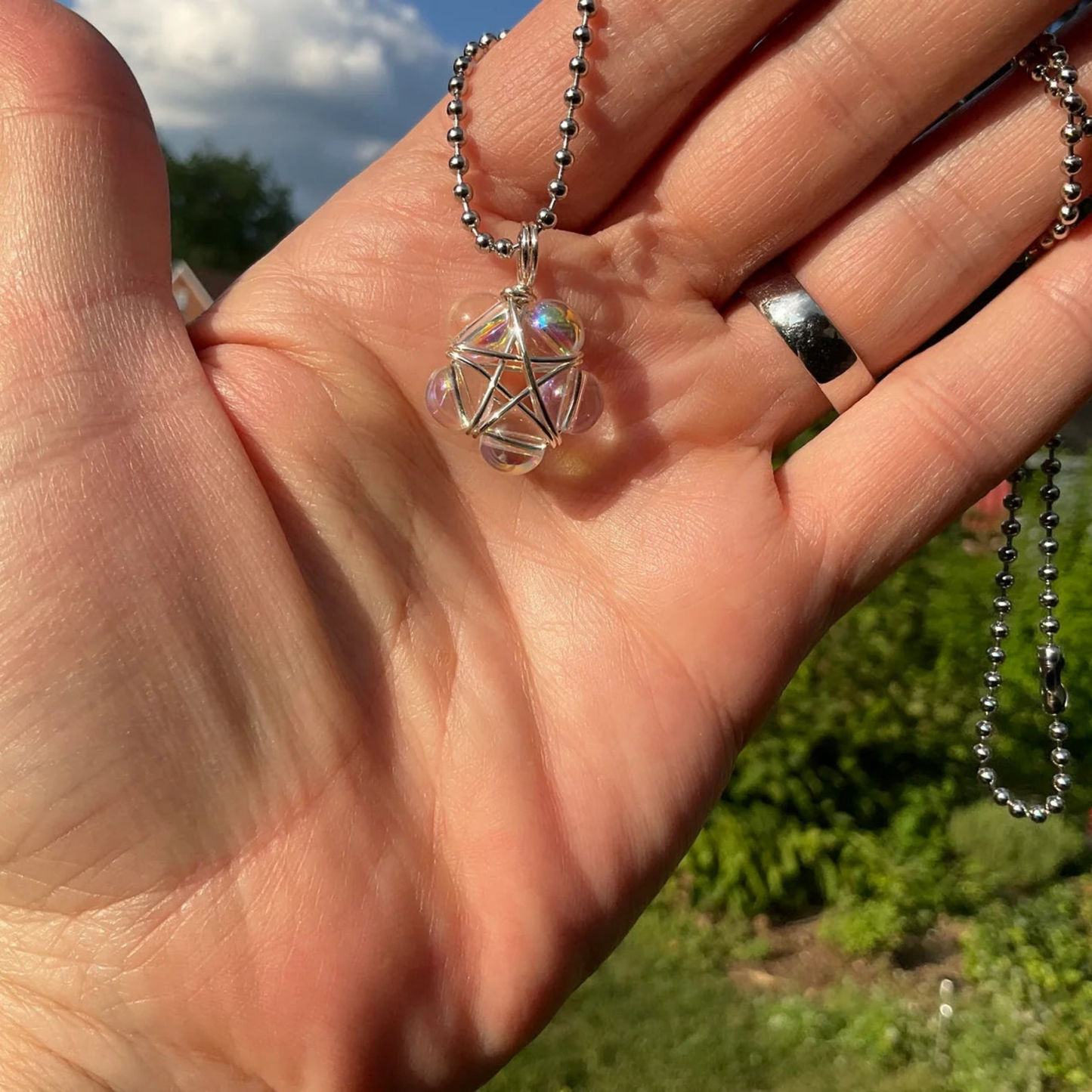 Hand holding a ball chain necklace with a bubble bead wrap pentagram pendant against a natural garden background