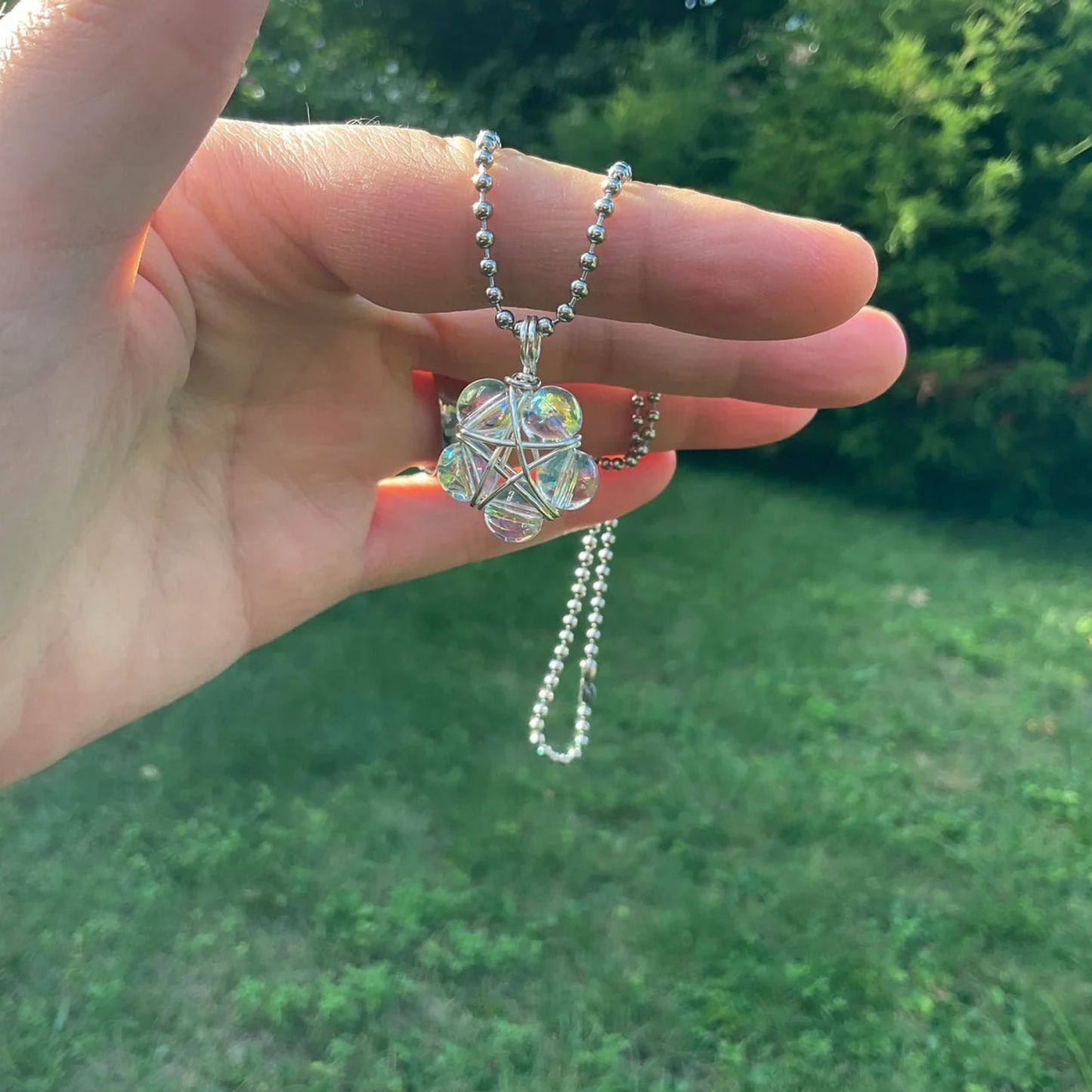 Hand holding a silver tone ball chain necklace with an iridescent bubble bead wire wrap Pentagram pendant against a green garden outdoor background