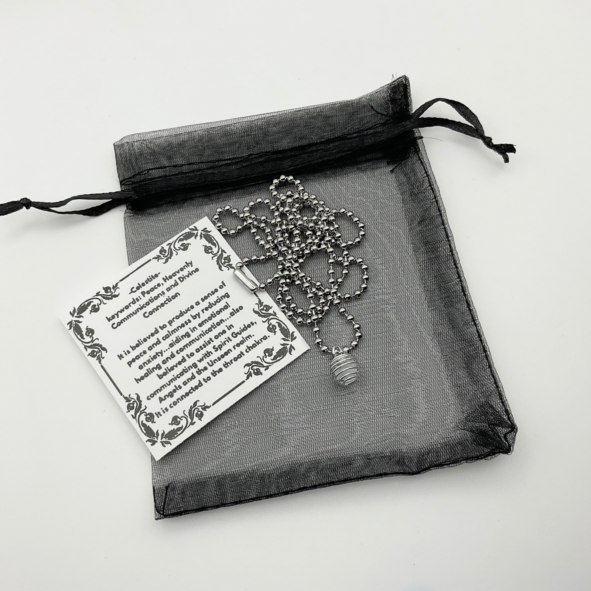 Necklace with silver tone celestite chip cage pendant and metaphysical properties stickerin a black organza drawstring bag on a light background