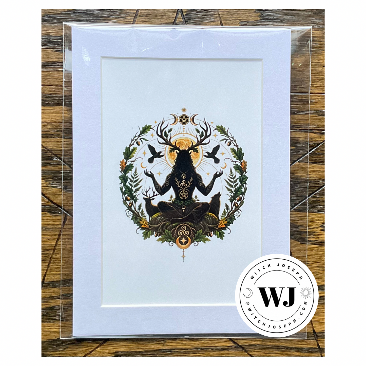 A art print featuring a black Cernunnos silhouette figure with deer antlers and a golden sun, surrounded by green foliage and birds, a wolf and a deer, in a white mat, presented in a clear protective sleeve on a rustic wood background.  The Witch Joseph logo is visible in the right hand lower corner.