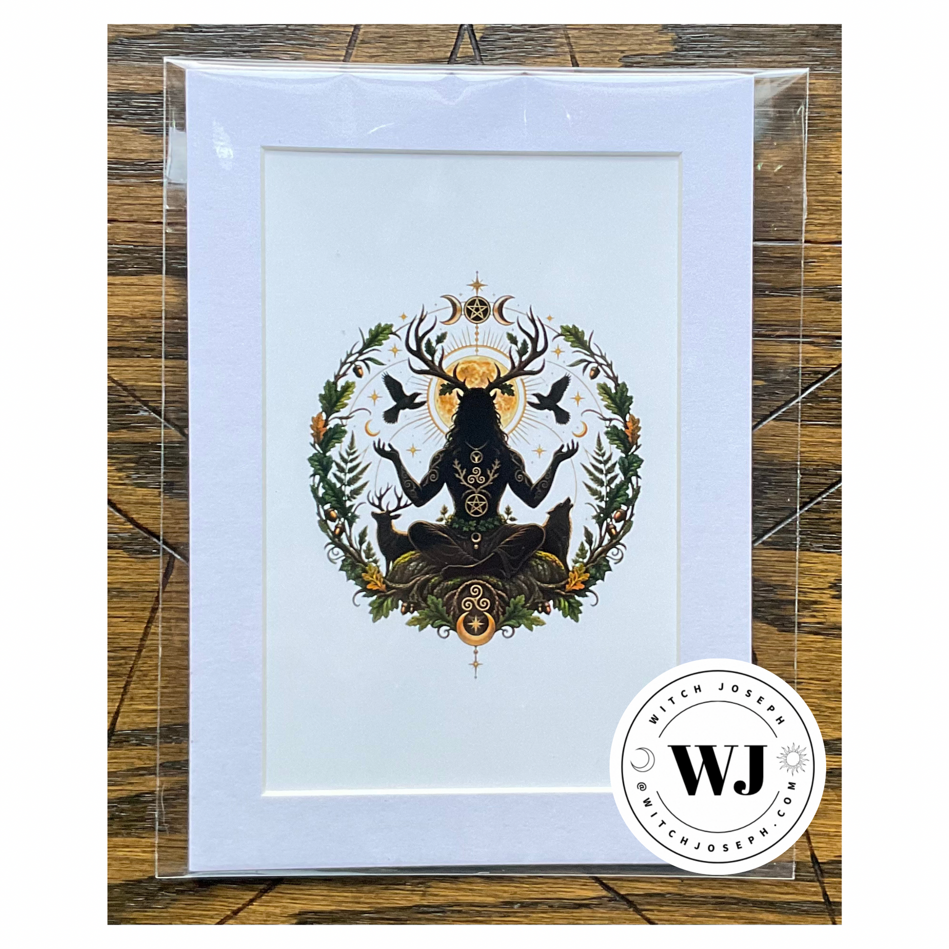 A art print featuring a black Cernunnos silhouette figure with deer antlers and a golden sun, surrounded by green foliage and birds, a wolf and a deer, in a white mat, presented in a clear protective sleeve on a rustic wood background.  The Witch Joseph logo is visible in the right hand lower corner.