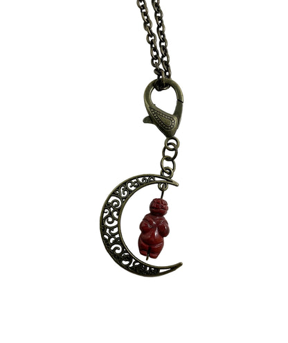A metal pendant necklace featuring a crescent moon with ornate filigree detailing and a small red jasper Goddess carved figurine hanging in the center, suspended from a chain with a lobster clasp, shown on a plain white background.