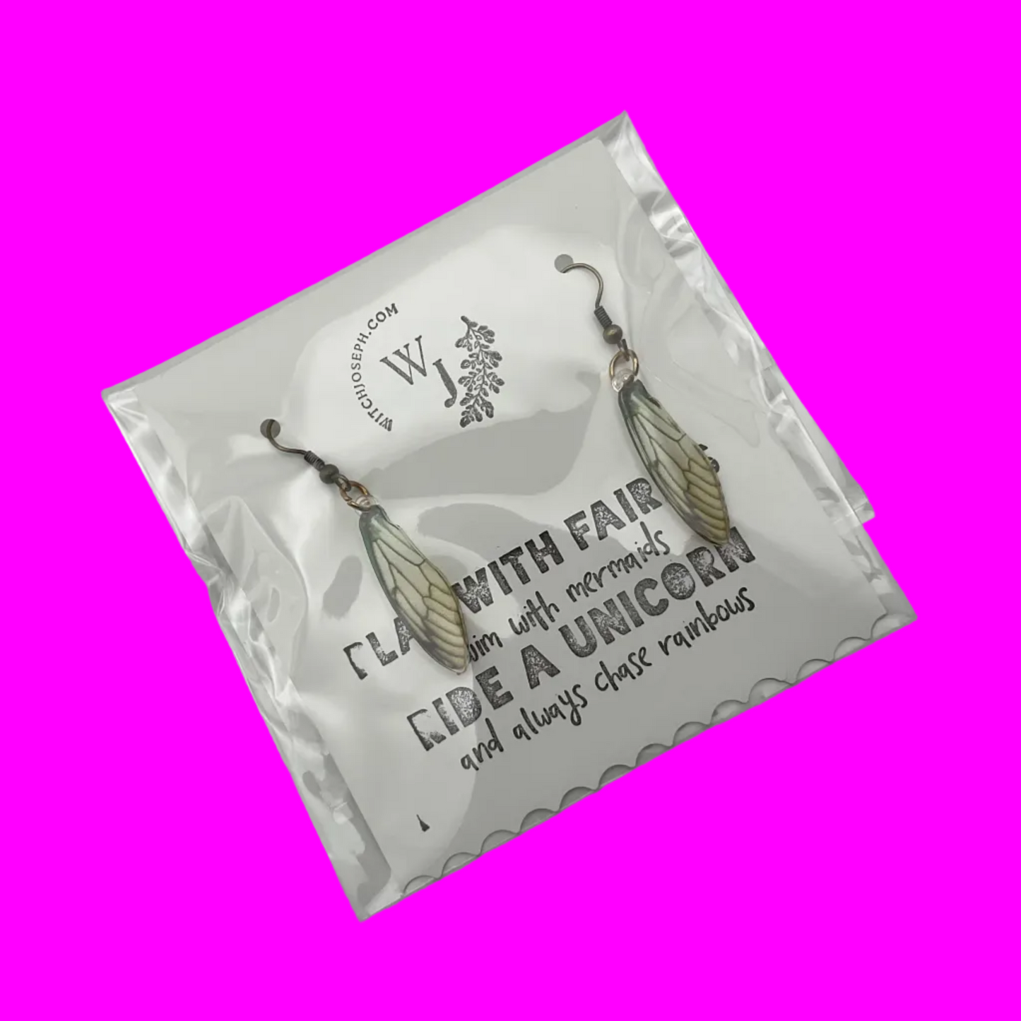Fairy Wing Earrings shown in whimsical packaging with text that says “Play with Fairies, swim with mermaids, Ride a Unicorn…and always chase rainbows.” Shown against a neon pink background