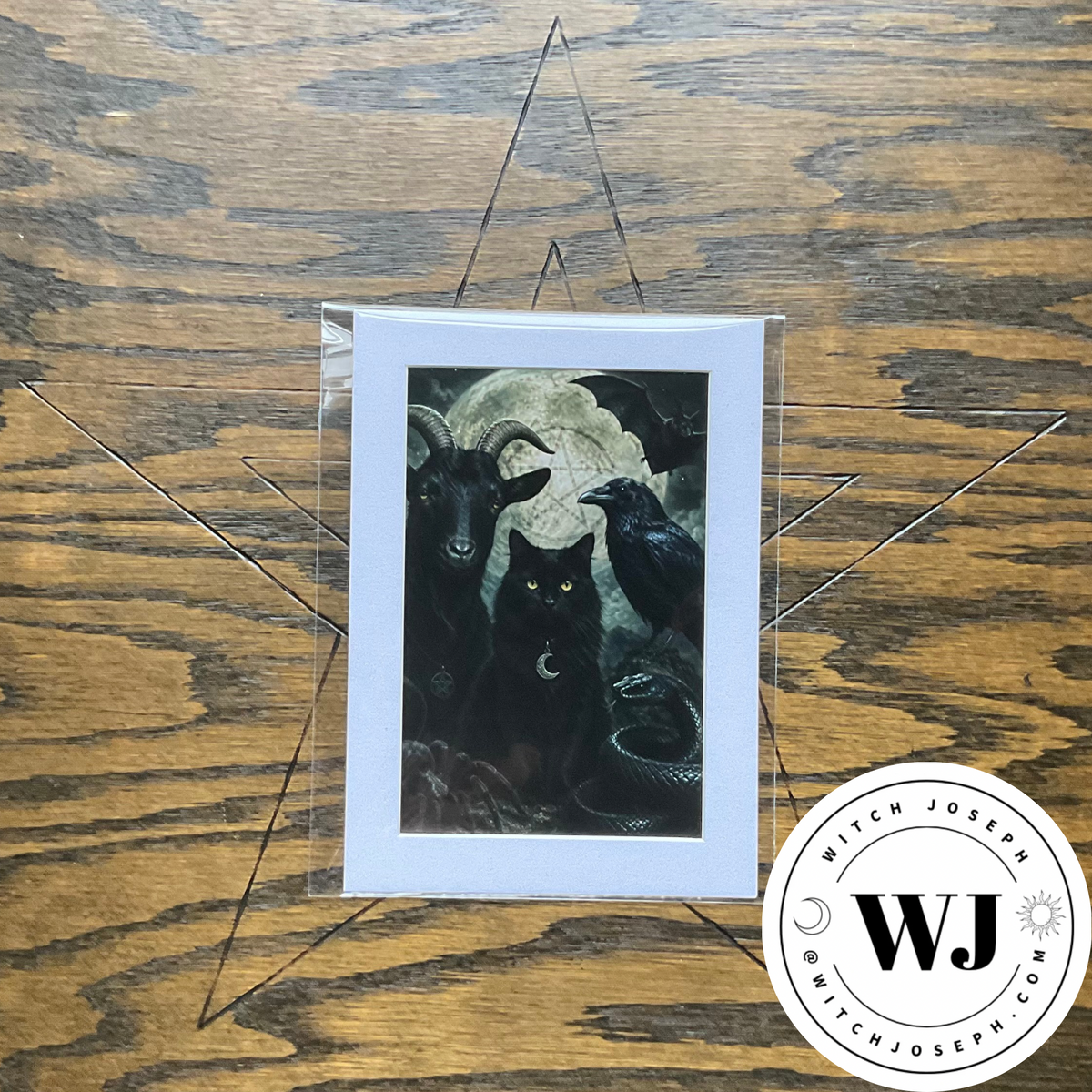 A Familiar spirits art print in mat featuring a dark illustration of a black cat, goat, crow, and snake and other creatures in front of a full moon, displayed on a wooden surface with a clear protective sleeve. A circular 'Witch Joseph' logo watermark is visible in the bottom right corner.